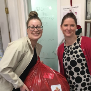Two women standing together, smiling, with one holding a large red donation bag. The woman on the left is wearing glasses and a light-colored jacket, while the woman on the right is wearing a black and white polka dot dress with a red cardigan. Behind them is a door with a sign that says 'Welcome to the Children's Cottage' and a wall with various posters and decorations.