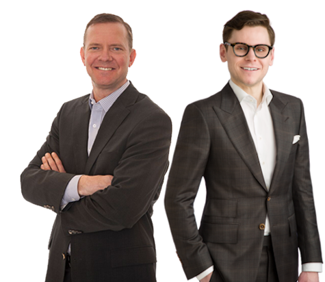 A portrait of two men from Scott Land & Lease Ltd. From left to right: Greg Meidinger, VP Land, smiling and wearing a dark suit with his arms crossed; and Hunter Scott, Executive Vice President, wearing glasses and a dark suit with a checkered shirt, smiling with his hands in his pockets. Both are standing against a white background, conveying a sense of professionalism and confidence.