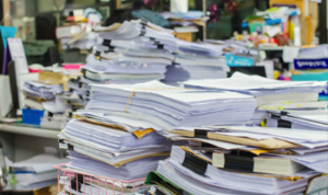 A cluttered office desk piled high with numerous stacks of paperwork, documents, and files. The papers are in various states of organization, with some neatly stacked and others scattered haphazardly. The background shows a busy office environment with more stacks of papers, office supplies, and equipment, creating a sense of overwhelming workload and disorganization.