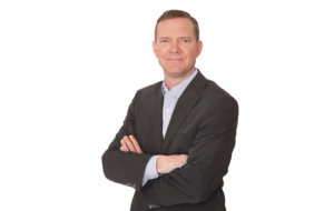 A portrait of Greg Meidinger, VP Land at Scott Land & Lease Ltd. He is wearing a dark suit with a light-colored shirt and is standing with his arms crossed, looking directly at the camera with a neutral expression. The background is plain white, highlighting his professional appearance.