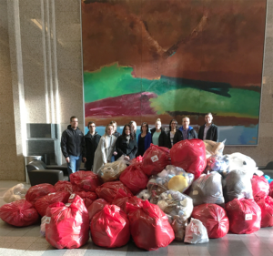 A group of Scott Land & Lease team members standing in front of a large pile of red and white bags filled with items, likely for donation. The group is indoors, in a spacious area with high ceilings and a large, colorful abstract painting on the wall behind them. The team members are smiling, dressed in business casual attire, and appear to be participating in a charitable activity.