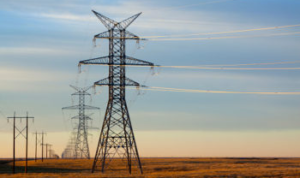 Several high-voltage power transmission towers stretching into the distance across an open landscape. The towers are connected by multiple power lines, set against a backdrop of a clear sky with subtle clouds. The scene illustrates the infrastructure used for electricity transmission over long distances.